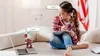 A young girl is sitting on a couch, writing in a notebook, while interacting with a small robot on a laptop. Náhľad obrázku produktu