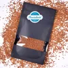 A black resealable pouch filled with brown sprinkles, placed on a white surface scattered with more sprinkles. Produktbild-Vorschau