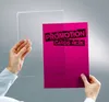 Hands holding a clear acrylic sheet over a pink promotional card with text. Anteprima dell’immagine prodotto