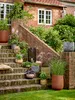 A collection of decorative pots with plants arranged on brick steps leading up to a house. Produktbild-Vorschau