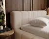 A close-up view of a textured boucle upholstered bed frame with a white pillow on it, set against a wooden paneled wall. Náhled obrázku produktu