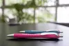 Two pens on a dark surface with a blurred background of greenery and windows. Produktbild-Vorschau