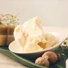 A close-up of shea butter in a white bowl, with soap bars and dried flowers in the background. Produktbild-Vorschau