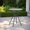 A round bistro table with a dark gray metal frame is placed on a light gray paved patio with green bushes and trees in the background. Produktbild-Vorschau