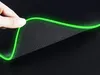A hand is shown lifting the edge of a black mousepad with a glowing green edge. Produktbild-Vorschau