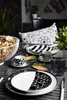A table setting with black and white patterned dishes, cups, and glasses, along with a bowl of pretzels and a decorative pillow. Produktbild-Vorschau