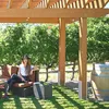A woman sits on a couch under a wooden pergola, with a vineyard in the background and a wine barrel to the side. Produktbild-Vorschau