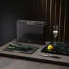 A set of black cutting boards with a holder, along with kale, a lemon, a glass of champagne, and a knife, arranged on a wooden surface. Anteprima dell’immagine prodotto