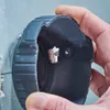 A hand is holding a black circular tool with a silver metal insert against a light gray wall. Produktbild-Vorschau