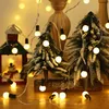 Christmas lights are draped around miniature snow-covered Christmas trees and a small decorative house with a snowman inside. Náhled obrázku produktu