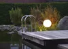 A garden scene at dusk with a wooden deck, pond, and a glowing spherical garden light. Produktbild-Vorschau
