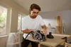 A man is using a circular saw to cut a piece of wood in a workshop or room with windows. Produktbild-Vorschau