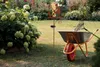 A garden scene with a solar-powered rain gauge and a wheelbarrow filled with gardening tools. Anteprima dell’immagine prodotto