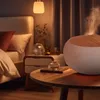 A humidifier is shown on a wooden table next to a bed in a bedroom. Podgląd zdjęcia produktu