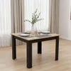 A square dining table with a light wood top and black legs, set with placemats, plates, and cutlery. A vase with greenery and a water bottle are on the table. The background features light-colored curtains. Voir les images du produit