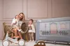 A mother is holding a baby while a toddler stands next to a playpen and a balance bike. Náhľad obrázku produktu