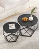 Two nesting round coffee tables with black wood tops and geometric black metal bases are placed on a textured rug next to a white sofa. One table has a book and coffee cup, the other has a bowl of oranges and a vase with a green stem. Náhled obrázku produktu