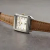 A close-up shot of a square-faced women's watch with a brown leather strap, resting on a textured gray surface. Podgląd zdjęcia produktu