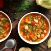 Two bowls of vegetable soup on a wooden table, with a slice of bread and herbs. Podgląd zdjęcia produktu