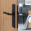 A black door handle set on a wooden door, with a bedroom scene in the background. Produktbild-Vorschau