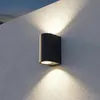 A dark gray, cylindrical outdoor wall light is mounted on a textured white wall, casting light upwards and downwards. Náhled obrázku produktu
