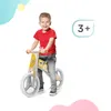 A young boy is riding a wooden balance bike. The background has abstract shapes in light blue, yellow, and pink. Náhľad obrázku produktu