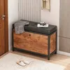 A shoe cabinet with a padded seat, placed in an entryway next to a door and a wall with vertical paneling. A blanket and a camera are on the seat, and slippers are on the floor in front of it. Podgląd zdjęcia produktu