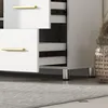 A close-up view of a white bathroom cabinet with gold handles and silver legs, placed on a light gray rug. Produktbild-Vorschau