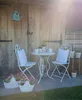 A charming outdoor seating area with a small table and chairs set up for tea, decorated with potted plants and a birdhouse. Podgląd zdjęcia produktu