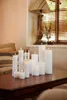 A collection of white LED candles of varying heights are arranged on a dark wooden table in a softly lit room. Produktbild-Vorschau