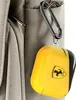 A yellow Ferrari-branded AirPods case is attached to a carabiner, which is clipped to a pocket on a gray bag. Náhľad obrázku produktu