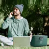 A man is sitting on the grass in a park, talking on his phone and working on his laptop. A cooler box and a travel mug are next to him. Produktbild-Vorschau