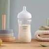 A Philips Avent glass baby bottle filled with milk sits on a wooden surface, next to a stack of folded towels and a small spoon. Podgląd zdjęcia produktu