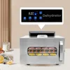 A stainless steel food dehydrator with food inside, placed on a table with a candle and fruit nearby. A callout box highlights the control panel. Podgląd zdjęcia produktu
