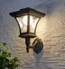 A black outdoor wall lantern with multiple bulbs is mounted on a textured white brick wall. A green plant is visible in the lower-left corner. Produktbild-Vorschau
