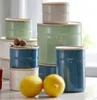 A collection of ceramic storage jars with wooden lids, some lemons, and nuts on a countertop. Produktbild-Vorschau