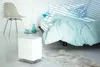 A modern bedroom with a white humidifier in the foreground and a bed with light blue bedding in the background. Podgląd zdjęcia produktu