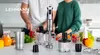 A kitchen scene with a blender and other kitchen accessories on a counter, with two people in the background. Produktbild-Vorschau