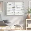 A white display cabinet with multiple shelves is mounted on a light gray wall. The cabinet is empty except for a few small decorative items on some shelves. In the foreground, a gray armchair and a gray ottoman are placed on a rug. Náhľad obrázku produktu