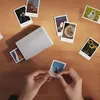 A person's hands are peeling a photo from a sticker backing, surrounded by other printed photos and a portable photo printer on a wooden table. Anteprima dell’immagine prodotto
