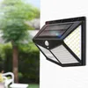 A solar-powered outdoor light with motion sensor is mounted on a white wall, with a blurred background of trees and greenery. Voir les images du produit