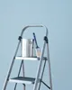 A silver stepladder with two steps, a paint can, and a paintbrush on the top step, against a light blue background. Náhľad obrázku produktu