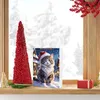 A Christmas-themed advent calendar featuring a cat in a Santa hat is displayed on a shelf next to a decorative red tree and a gift box, with a snowy window in the background. Produktbild-Vorschau