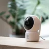 A white Sinji pan-tilt camera is placed on a wooden surface with a blurred green plant in the background. Produktbild-Vorschau