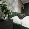 A close-up view of a bedroom with a green upholstered bed, white bedding, and a dark bedside table with a vase of white flowers. Náhled obrázku produktu