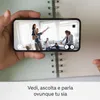 A hand holds a smartphone displaying a live video feed of a family in a living room, superimposed over an open notebook. Produktbild-Vorschau