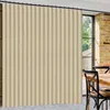 A beige vertical blind is shown covering a large window in a room with exposed brick walls and a wooden table with chairs. Produktbild-Vorschau