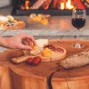A hand is picking up a piece of cheese from a cheese board in front of a fireplace. A glass of red wine and a baguette are also on the table. Produktbild-Vorschau