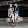 Two women standing in a modern, dimly lit urban setting, each holding a duffel bag. Anteprima dell’immagine prodotto