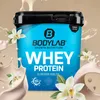 A tub of BODYLAB Whey Protein with splashes of milk and vanilla flowers. Produktbild-Vorschau